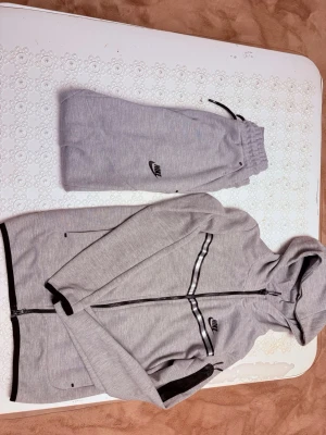 Gråa Nike tech set - Snygga gråa mjukisbyxor från Nike med svart logga på benet. Byxorna har resår i midjan med svart dragsko och muddar vid bensluten. Tillverkade i mjukt och bekvämt material, perfekta för chill dagar.