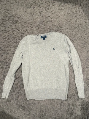 Ljusgrå tröja från Polo Ralph Lauren - Klassisk ljusgrå tröja från Polo Ralph Lauren med rund halsringning och den ikoniska broderade loggan på bröstet. Tröjan har långa ärmar och ribbade muddar vid ärmslut och nederkant. Perfekt för en stilren och enkel look.