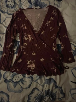 Blommig omlottopp i vinrött XS - Säljer en vinröd omlottopp med blommigt mönster och långa ärmar. Toppen har v-ringning och knyt i sidan, vilket ger en feminin och trendig look. Perfekt för dig som gillar romantiska detaljer och mjuka material.