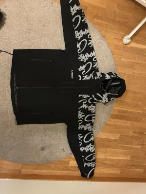 Corteiz windbreaker  - Snygg svart vindjacka med grått graffiti-inspirerat mönster på ärmar och huva. Jackan har dragkedja framtill, två sidofickor med dragkedja och justerbar huva. Perfekt för dig som vill sticka ut med en unik streetwear-look.