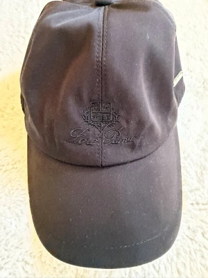Svart keps från Loro Piana - Stilren svart keps från Loro Piana med broderad logga framtill och en liten metallbricka på sidan. Klassisk böjd skärm och justerbar passform. Materialet ser ut att vara mjukt och vattenavvisande, perfekt för en clean och sportig look.