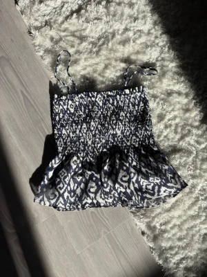 Söt topp - Blåvit topp med volang nedtill. Toppen har tunna axelband och ett snyggt grafiskt mönster. Perfekt för sommardagar och enkel att matcha med jeans eller kjol. Man kan även ta av banden och göra den till en tubetopp