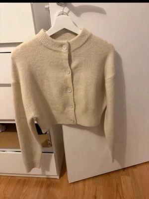 Beige croppad kofta med knappar - Mysig croppad kofta i beige med rund hals och stora matchande knappar framtill. Tröjan har långa ärmar och ribbade muddar vid hals, ärmslut och nederkant. Materialet ser fluffigt och mjukt ut, perfekt för lager på lager.