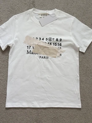 Vit t-shirt med tryck från Maison Margiela - Vit t-shirt från Maison Margiela med svart tryck framtill som visar siffror och texten 'Maison Margiela PARIS'. Priset går att diskutera.