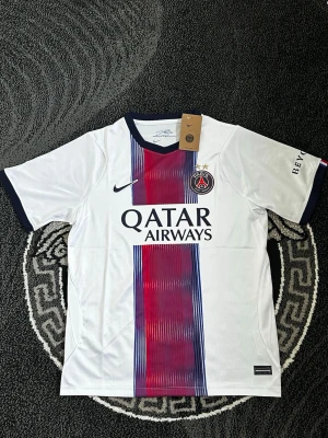 PSG vit matchtröja Nike - Snygg vit PSG matchtröja från Nike med mörkblå och röda vertikala ränder i mitten. Tröjan har korta ärmar, mörkblå detaljer vid krage och ärmslut samt PSG-logga och Qatar Airways-tryck på bröstet. Perfekt för fotbollsfans!