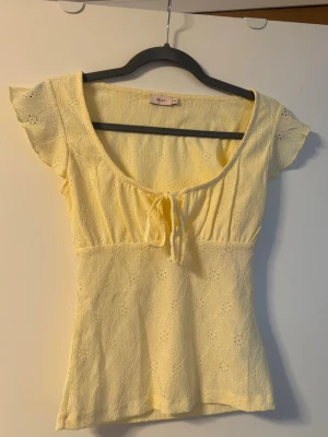 Söt gul topp från Nelly - Söt ljusgul topp från Nelly med korta ärmar och knyt framtill. Toppen har broderade detaljer i tyget och en feminin, lätt utsvängd passform. Perfekt för varma dagar och ger en fräsch look till din outfit. Helt ny med prislappen kvar