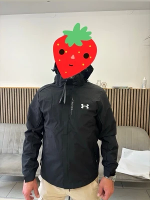 Svart vindjacka från Under Armour - Snygg svart vindjacka från Under Armour med dragkedja framtill, justerbar huva och två sidofickor med dragkedja. Jackan har en stilren logga på bröstet och är tillverkad i ett lätt, vattenavvisande material som passar perfekt för blåsiga dagar. Ny i förpackning 