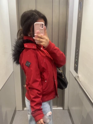 Röd Hollister jacka från 2000 talet - Snygg röd bomberjacka från Hollister med svart fuskpäls på huvan. Jackan har ribbade muddar, dragkedja och knappar framtill samt två fickor. Perfekt för höst och vinter när du vill hålla dig varm och se cool ut.