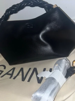 Svart handväska med flätat handtag Ganni - Snygg svart handväska från Ganni i veganskt läder med ett unikt flätat handtag. Väskan har en modern, kantig form och diskret logga framtill. Perfekt accessoar för att lyfta din outfit. Dustbag och axelrem medföljer. (Den behövs inte längre, har inte kommit till användning helt enkelt) ni kan buda