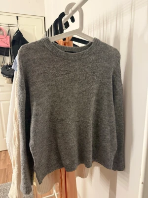 Grå stickad tröja från 157 - Mysig grå stickad tröja från 157 med rund halsringning och ribbade muddar vid ärmslut och nederkant. Enkel och stilren design som passar perfekt till jeans eller kjol. Tillverkad i mjukt material för extra komfort.