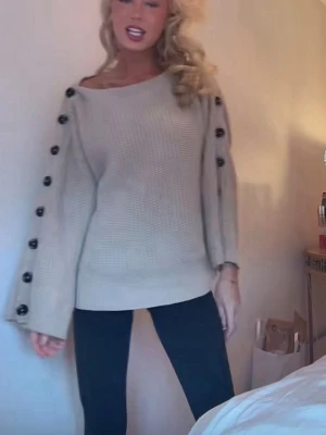 Beige stickad tröja med knappar - Beige stickad tröja med breda ärmar och svarta dekorativa knappar längs hela ärmsidorna. Tröjan har en lös passform och rund halsringning. Perfekt för dig som gillar en avslappnad och trendig stil.