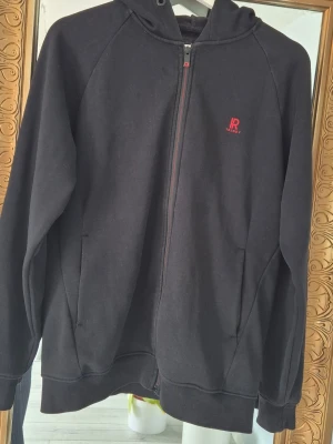 Svart hoodie med dragkedja - Svart hoodie med dragkedja och huva. Liten röd logga på bröstet med texten 'R READY'. Tillverkad i mjukt sweatshirtmaterial och har två fickor framtill. Perfekt för en avslappnad och stilren look.