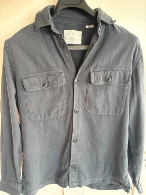 Mörkgrå overshirt med fickor - Stilren mörkgrå overshirt i mjukt bomullsmaterial. Skjortan har två stora bröstfickor med lock och knapp, klassisk krage och knäppning framtill. Perfekt lager-på-lager-plagg för en avslappnad look.