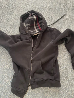 Svart hoodie med rutigt foder - Säljer en svart hoodie med dragkedja och huva. Insidan av huvan har ett rutigt foder i grått, vitt och rött. Hoodien har fickor framtill och ribbade muddar vid ärmar och nederkant. Perfekt för en avslappnad stil. Själva saken man drar upp dragkedjan med har gått av som visas på sista bilden men dragkedjan fungerar fortfarande 