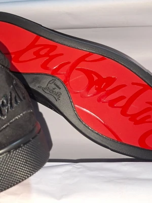 Svarta Louboutin sneakers med röd sula - Snygga svarta sneakers från Christian Louboutin i mocka med klassisk röd sula och svarta skosnören. Låg modell med diskret logga på sidan och insidan. Perfekta för dig som vill ha en exklusiv touch på din outfit.
