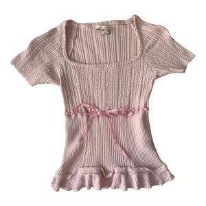 Kimchi blue Ljusrosa ribbad topp med rosett - Supersöt ljusrosa topp med ribbad struktur, korta ärmar och volangkant nedtill. Toppen har en dekorativ rosett i midjan och en fyrkantig ringning. Perfekt för dig som gillar romantisk och feminin stil.