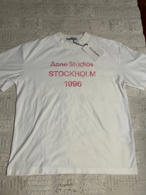 Vit Acne Studios t-shirt Stockholm - Vit t-shirt från Acne Studios med rosa text 'Acne Studios STOCKHOLM 1996' på bröstet. Klassisk passform och rund hals. Perfekt för dig som gillar stilrena plagg med en twist.