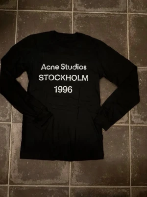Svart långärmad topp Acne Studios - Svart långärmad topp från Acne Studios med vit text framtill där det står 'Acne Studios STOCKHOLM 1996'. Klassisk rund halsringning och rak passform. Perfekt för dig som gillar stilrena och trendiga plagg. Är ca 182 och väger 66