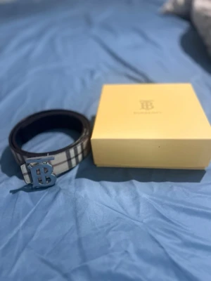 Burberry bälte - Snyggt bälte med klassiskt randigt mönster i svart, vitt och beige. Bältet har ett stort silvrigt spänne med initialerna 'TB' framtill. Perfekt accessoar för att lyfta din outfit.