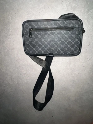 Svart axelväska med monogrammönster - Snygg svart axelväska med justerbar rem och monogrammönster över hela väskan. Väskan har en dragkedjeficka på framsidan och en stilren, rektangulär form. Materialet ser ut att vara slitstark canvas med detaljer i konstläder.