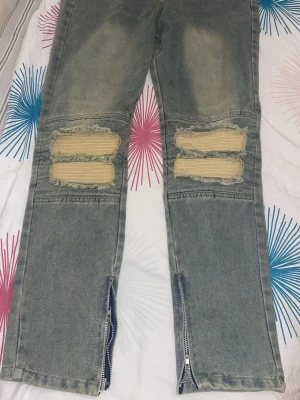 Grå slitna jeans med dragkedjor - Säljer ett par grå jeans med slitningar och hål över knäna, samt dragkedjor vid bensluten. Jeansen har en rak passform och tydliga slitdetaljer för en trendig look. Perfekta för dig som gillar streetwear och vill sticka ut.