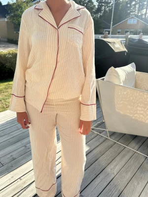  Lxa pyjamas  - Klassisk tvådelad pyjamas i beige med tunna vita ränder och röda passpoaler. Skjortan har krage, bröstficka och knappar framtill. Byxorna har raka ben och ficka bak. Jag har Xs i tröjan och S i byxorna. Pyjamasen är köpt på retuned och prislapp finns kvar på byxorna. 