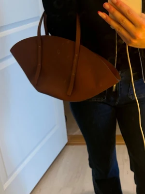 Brun handväska i läderlook - Snygg brun handväska med unik bågformad design och två långa axelband. Väskan är gjord i ett slätt, läderliknande material och har en minimalistisk look utan synliga loggor eller detaljer. Perfekt för dig som gillar stilrena accessoarer. Aldrig använd 