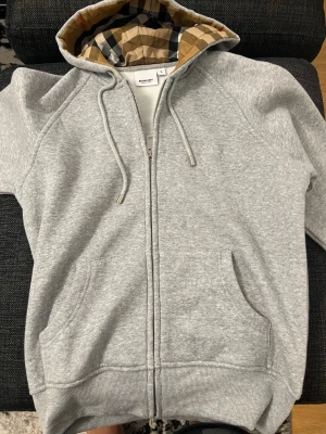 Grå zip hoodie från Burberry - Snygg grå zip hoodie från Burberry med klassiskt rutigt foder i huvan. Hoodien har dragsko i huvan, två fickor framtill och ribbade muddar. Tillverkad i mjukt sweatshirtmaterial som är perfekt för en chill och stilren look.