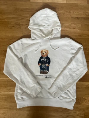 Vit hoodie Polo Bear Ralph Lauren - Vit hoodie med huva och dragsnören från Ralph Lauren. På bröstet finns ett tryck av den ikoniska Polo Bear i slitiga blå tröja och jeans. Under björnen står texten 'POLO BEAR BY RALPH LAUREN'. Perfekt för en avslappnad och trendig stil. Skriv gärna om ni har frågor mm, priset kan diskuteras vid rimliga bud.