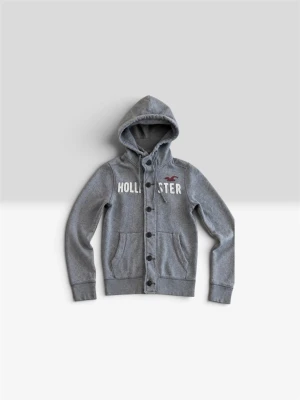 Grå hoodie från Hollister  - Snygg grå hoodie från Hollister med knappar framtill och vit logga på bröstet. Tröjan har huva med dragsko och två fickor fram. Perfekt för en avslappnad och cool stil. Tillverkad i mjukt sweatshirtmaterial.