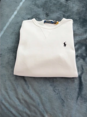 Vit sweatshirt från Polo Ralph Lauren - Klassisk vit sweatshirt från Polo Ralph Lauren med rund halsringning och broderad logga i svart på bröstet. Tröjan har ribbade muddar vid ärmslut och nederkant. Perfekt för en clean och stilren look.