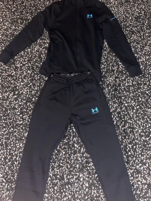 Under Armour Black Tracksuit Set - This is a black Under Armour tracksuit set, consisting of a zip-up hoodie and matching jogger pants. The set features the Under Armour logo in blue on both the jacket and pants. The waistband of the pants is branded with 'UNDER ARMOUR'. The tracksuit is laid out on a black and white patterned carpet, shown from both the front and back.
