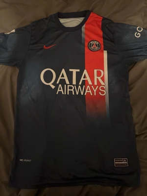 PSG matchtröja Messi #30 - Snygg mörkblå PSG matchtröja från Nike med röd och vit rand på framsidan, Qatar Airways-tryck och klubbmärke. På ryggen står det Messi och nummer 30. Perfekt för fotbollsfans som vill stötta sitt lag på match eller träning.