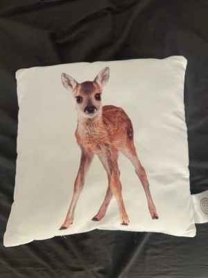 Vit kudde med rådjurmotiv - Säljer en fyrkantig vit kudde med ett realistiskt tryck av ett rådjur på framsidan. Kudden är mjuk och har en tydlig djurdesign som passar perfekt som inredningsdetalj i soffan eller sängen. Materialet känns lent och motivet är detaljerat.