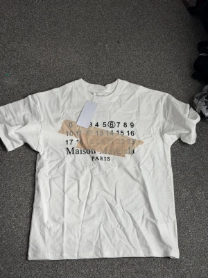 Maison Margiela vit t-shirt - Vit t-shirt från Maison Margiela med ikoniskt siffertryck och beige tejpdetalj på bröstet. Klassisk passform och rund hals. Perfekt för dig som gillar streetstyle och designerplagg.