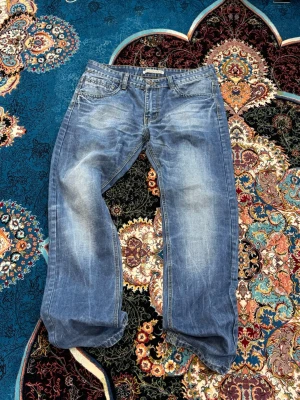 Blå raka jeans med slitningar - Säljer ett par blå jeans med raka ben och snygga slitningar på framsidan. Jeansen har klassisk femficksdesign och ljus tvätt som ger en avslappnad look. Perfekta för dig som gillar en casual stil.