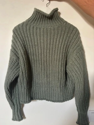 Grågrön stickad polotröja - Säljer en chunky stickad tröja i grågrönt med hög polokrage och breda ribbstickade muddar. Tröjan har en oversized passform och är perfekt för kyliga dagar. Materialet känns mjukt och mysigt, och modellen är lite boxig med långa ärmar.