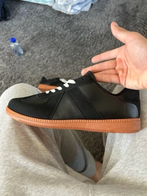 Maison Margiela Gats - Stilrena svarta sneakers med vit snörning och klassisk brun gummisula. Maison Margiela liknande. Skorna har en låg profil och är tillverkade i slätt material med diskreta sömmar för en clean look. Perfekta för dig som gillar minimalistisk stil. Saknar låda och skön inner sula men de går att fixa. Storleken var inte min men skulle säga att de passar 43-44.