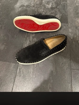 Svarta loafers med nitar och röd sula - Säljer ett par svarta loafers i mocka med coola svarta nitar runt hela skon. Skorna har en vit sula och en ikonisk röd undersida. Slip-on modell som ger en edgy och trendig look. Perfekta för dig som vill sticka ut med din stil. OBS FINNS INGE INNESULA
