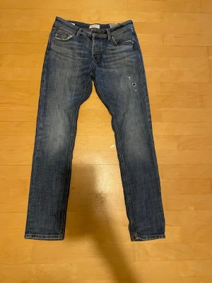 Blå raka jeans med slitningar - Säljer ett par klassiska blå jeans med raka ben och lätt slitna detaljer på framsidan. Jeansen har fem fickor och normal midja. Perfekta för en avslappnad och trendig look. Materialet är denim i bomull.