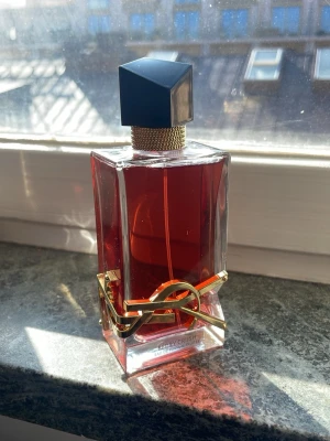 Helt ny Lyxig parfymflaska med gulddekor 50 ml  - Snygg parfymflaska med mörkröd vätska och en unik, geometrisk svart kork. Flaskan är rektangulär med en elegant gulddetalj runt om, vilket ger en exklusiv känsla. Perfekt för dig som vill ha en statement-doft på hyllan.