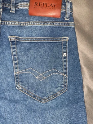 Blå jeans från Replay - Säljer ett par klassiska blå jeans från Replay med snygg slitning och raka ben. Jeansen har fem fickor, tydliga sömmar och en läderpatch med Replay-loggan bak i midjan. Materialet är robust denim som passar perfekt till vardagslooken. Kan sänka priset vid snabb affär.