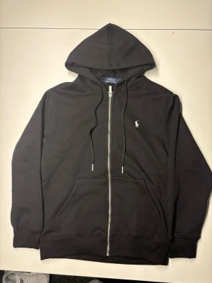 Svart zip-hoodie från Polo Ralph Lauren - Snygg svart hoodie med dragkedja från Polo Ralph Lauren. Klassisk design med huva, dragsnören och två fickor framtill. Liten vit logga broderad på bröstet. Tillverkad i mjukt bomullsmaterial som är skönt att bära.