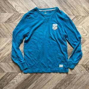 Blå långärmad tröja Tommy Hilfiger - Snygg blå långärmad tröja från Tommy Hilfiger med rund halsringning och broderad logga på bröstet. Tröjan har ribbade muddar och en liten etikett nertill. Perfekt för en avslappnad och stilren look.