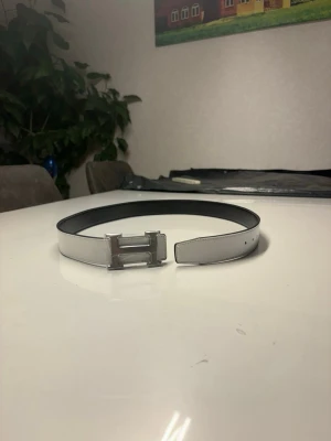 Vitt och svart bälte med metallspänne - Snyggt bälte i vitt med svart insida och ett stort metallspänne i form av ett H. Perfekt för att lyfta vilken outfit som helst med en clean och modern look. Bältet har en slät yta och klassisk design.