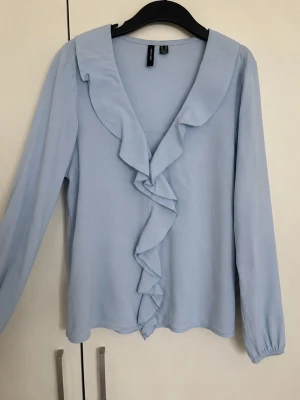 Ljusblå blus från Vera Moda - Superfin ljusblå blus från Vero Moda med fina volanger framtill. Den har långärmade ärmar och sitter väldigt fint på kroppen. Storlek XS.  Säljes eftersom jag inte använder den längre. Pris kan diskuteras!