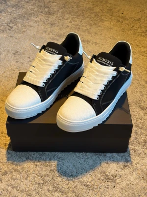 Svarta och vita sneakers från Numeris - Stilrena sneakers från Numeris i svart och vitt med chunky vit sula och breda vita skosnören. Ovandelen är i svart textil med vita detaljer och tåpartiet är i vitt läderliknande material. Snygg kontrastdesign och logga på plösen,Som kommer med extra snören.