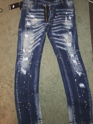 DSQUARED2 jeans mörk blå. Oanvänt jeans - Säljer ett par mörkblå jeans med slitningar, färgstänk och tvättade detaljer. Jeansen har raka ben, klassisk femficksmodell och markerade sömmar. Cool streetstil som sticker ut med både slitningar och färgprickar. Oanvänt. 