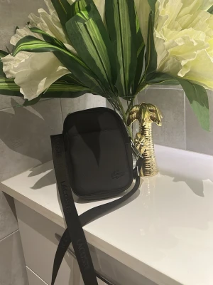 Svart axelväska från Lacoste  - Snygg svart axelväska från Lacoste med justerbar axelrem och diskret logga framtill. Väskan har en kompakt, rektangulär form och är tillverkad i slitstarkt material med dragkedja upptill och en extra ficka på sidan. Perfekt för att bära det viktigaste.