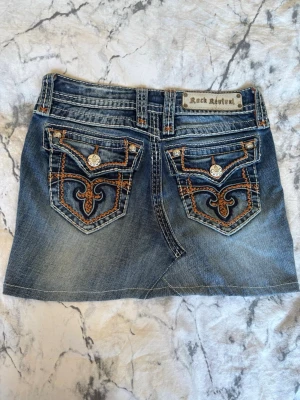Jeanskjol från Rock Revival - Snygg kort jeanskjol från Rock Revival med broderade bakfickor och dekorativa knappar. Kjolen har en klassisk blå denimfärg och detaljer i orange och vitt. Köptes från en secondhandbutik, men har aldrig kommit till användning av mig.💞 Kjolen har midjestorlek 25!